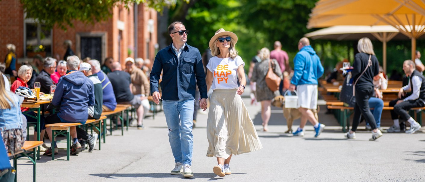 Ein Paar spaziert durch einen belebten Biergarten. Sie tragen lässige Kleidung und Sonnenbrillen. Im Hintergrund sitzen Menschen an Tischen unter Sonnenschirmen., © Katzenbacher Hof Ein Paar spaziert durch einen belebten Biergarten. Sie tragen lässige Kleidung und Sonnenbrillen. Im Hintergrund sitzen Menschen an Tischen unter Sonnenschirmen., © Katzenbacher Hof