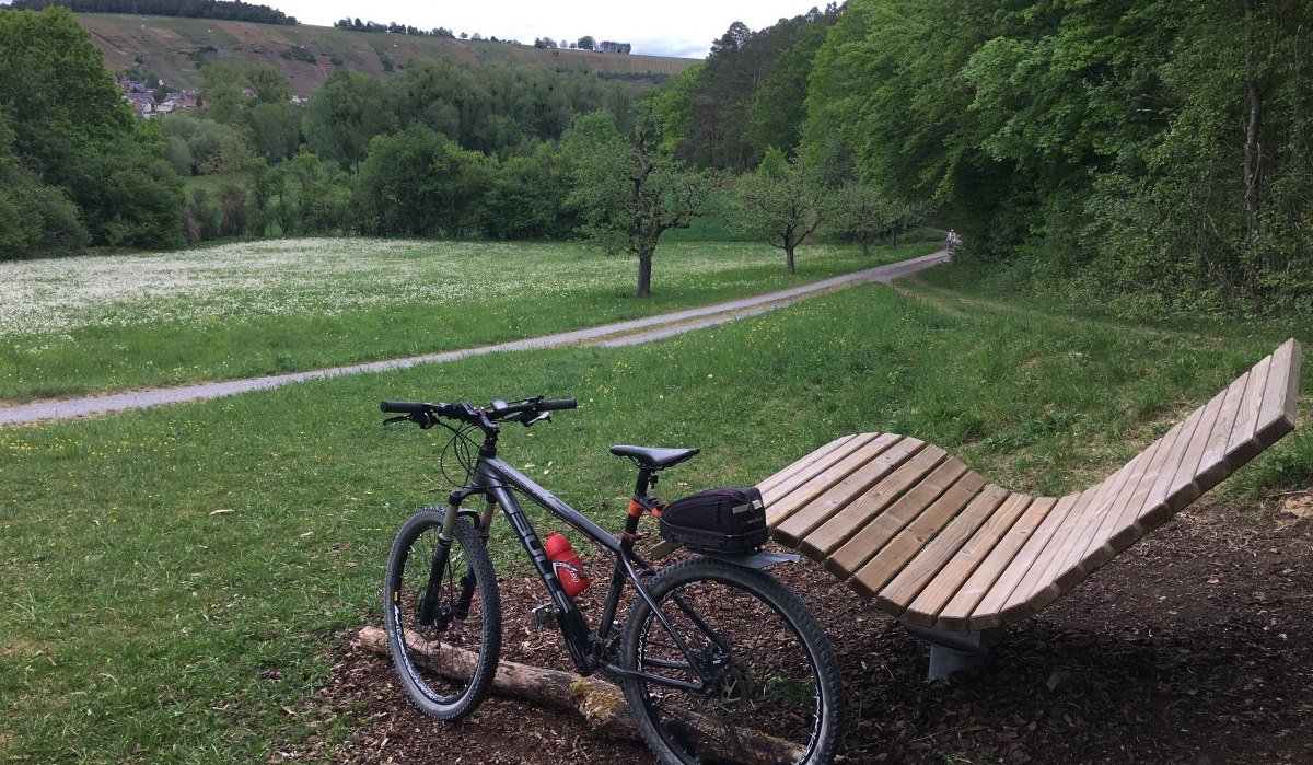 Fahrrad neben geschwungener Holzbank auf Wiese, umgeben von Bäumen und Hügeln im Hintergrund., © Land der 1000 Hügel - Kraichgau-Stromberg Fahrrad neben geschwungener Holzbank auf Wiese, umgeben von Bäumen und Hügeln im Hintergrund., © Land der 1000 Hügel - Kraichgau-Stromberg