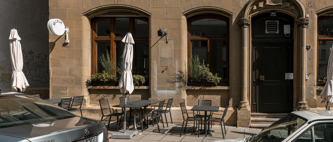 Gebäude mit Tischen und Stühlen auf dem Gehweg, Sonnenschirme und ein Schild mit 'MISIK'. Historische Fassade mit großen Fenstern., © Stuttgart-Marketing GmbH, Sarah Schmid