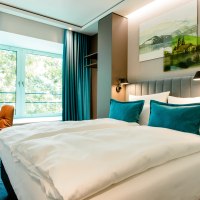 Modernes Hotelzimmer mit gro&szlig;em Bett, blauen Kissen und Vorh&auml;ngen. Ein Fenster bietet Blick ins Gr&uuml;ne. Ein Schreibtisch mit Stuhl steht daneben., &copy; Motel One - Stuttgart Hauptbahnhof