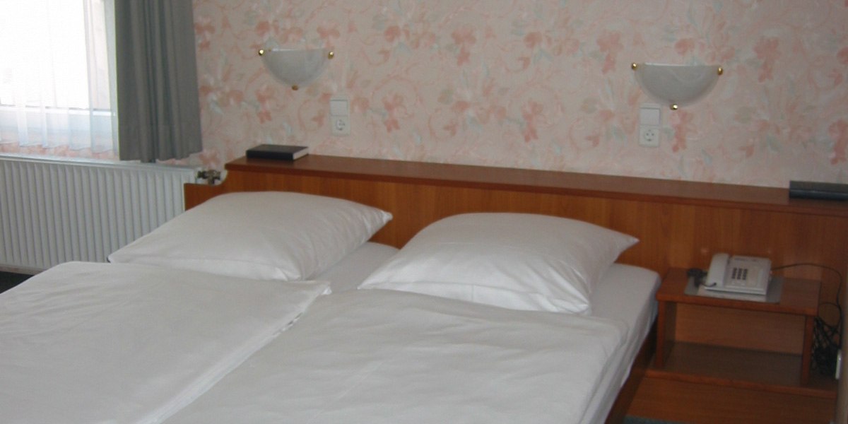 Ein Hotelzimmer mit zwei Einzelbetten, weißer Bettwäsche, blumiger Tapete und einem Nachttisch mit Telefon., © Hotel Wirt am Berg