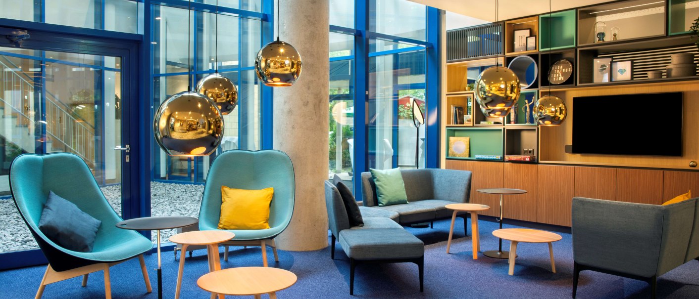 Moderne Lounge mit blauen Sesseln, Holztischen und goldenen Hängelampen. Große Fenster und Regale im Hintergrund schaffen eine einladende Atmosphäre., © Holiday Inn Stuttgart Moderne Lounge mit blauen Sesseln, Holztischen und goldenen Hängelampen. Große Fenster und Regale im Hintergrund schaffen eine einladende Atmosphäre., © Holiday Inn Stuttgart