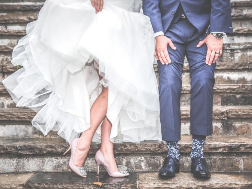 Ein Brautpaar sitzt auf einer Steintreppe. Die Braut tr&auml;gt ein wei&szlig;es Kleid und glitzernde High Heels, der Br&auml;utigam einen blauen Anzug mit gemusterten Socken., &copy; Canva