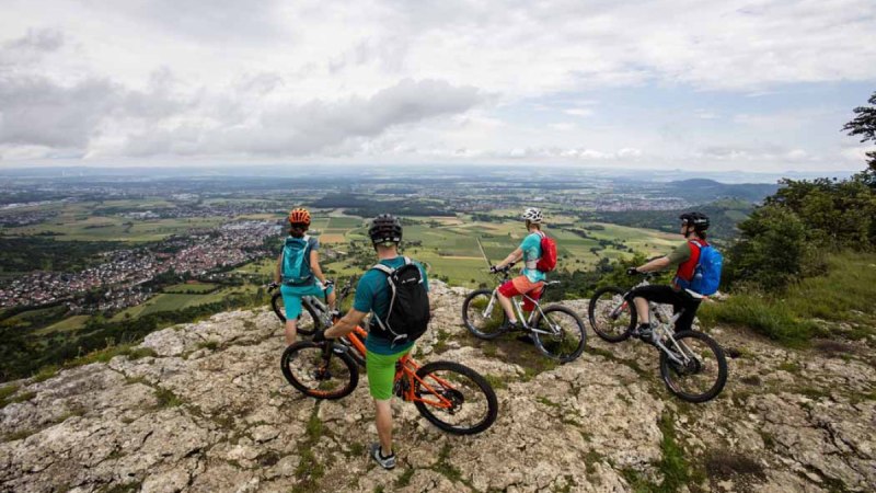 Vier Mountainbiker stehen auf einem Felsen und blicken auf eine weite Landschaft mit Feldern und D&ouml;rfern. Der Himmel ist bew&ouml;lkt., &copy; Schw&auml;bische Alb Tourismusverband e.V.
