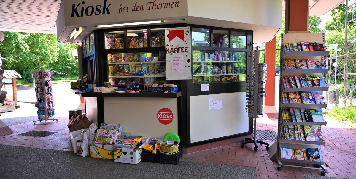 Ein Kiosk mit der Aufschrift 'Kiosk bei den Thermen', umgeben von Zeitschriftenständern und Verkaufsartikeln. Im Hintergrund sind Bäume sichtbar.