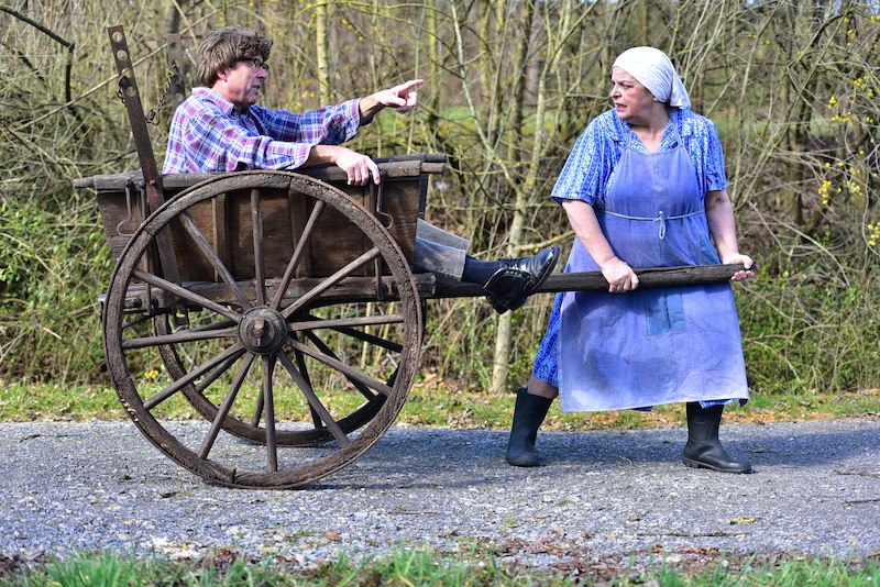 Eine Frau in Sch&uuml;rze zieht einen Mann in einem Holzwagen. Beide tragen l&auml;ndliche Kleidung und befinden sich auf einem Weg im Freien., &copy; EMT Event-Media-Tec GmbH