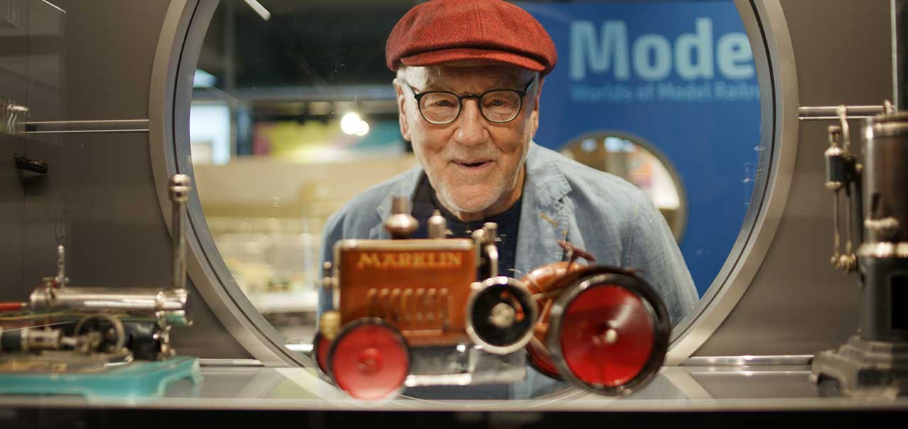 Ein älterer Mann mit rotem Hut blickt durch ein rundes Fenster auf ein Märklin-Modell eines Traktors in einer Ausstellung., © Märklineum