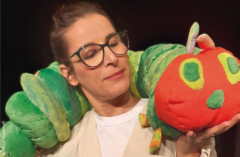 Frau mit Brille hält eine große Plüschraupe mit grünem Körper und rotem Kopf. Die Szene wirkt freundlich und kindgerecht., © Stadt Kornwestheim Kulturmanagement