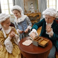 Drei Kinder in historischen Kostümen spielen Karten in einem prunkvollen Raum mit antiken Möbeln und Perücken., © Tourismus und Events Ludwigsburg