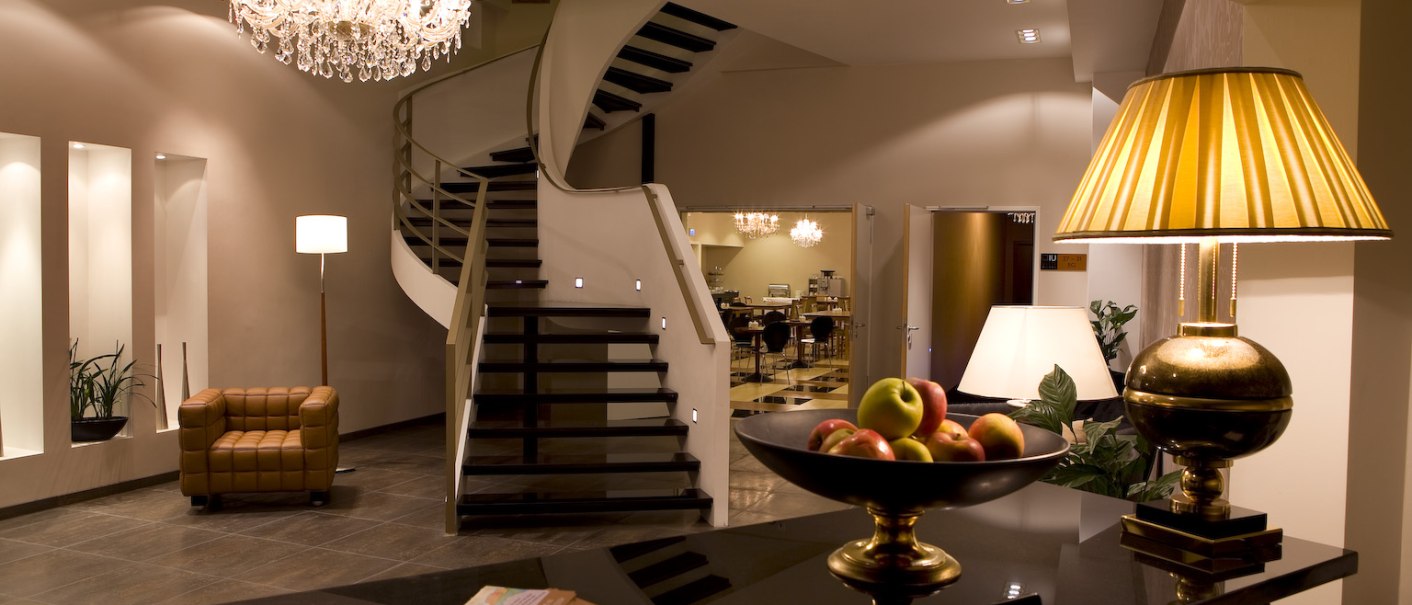 Elegante Hotellobby mit geschwungener Treppe, Kronleuchter, Tischlampe und Obstschale auf dem Tresen. Moderne Einrichtung mit Ledersessel., © Ross Messehotel Elegante Hotellobby mit geschwungener Treppe, Kronleuchter, Tischlampe und Obstschale auf dem Tresen. Moderne Einrichtung mit Ledersessel., © Ross Messehotel