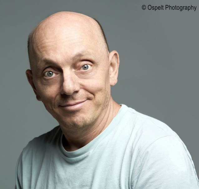 Ein Mann mit Glatze und hellem T-Shirt l&auml;chelt in die Kamera. Der Hintergrund ist grau und schlicht gehalten., &copy; Stadthalle Leonberg
