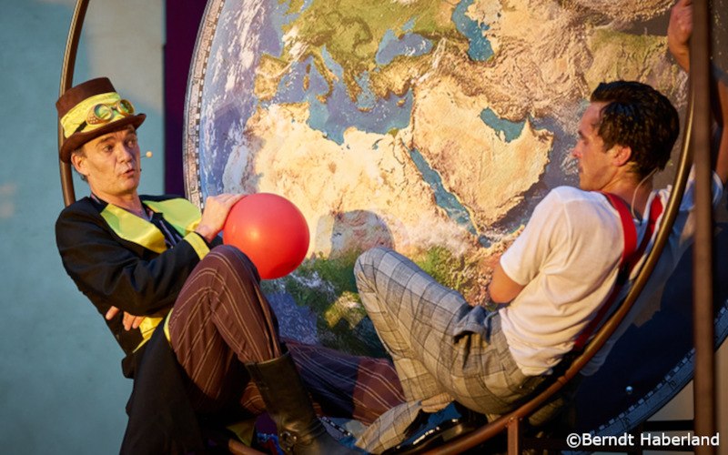 Zwei Männer in Kostümen sitzen vor einer großen Weltkarte. Einer trägt einen Zylinder und hält einen roten Ballon., © Kultur- und Sportamt Stadt Bietigheim-Bissingen