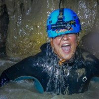Eine Person mit blauem Helm und Neoprenanzug lacht in einer nassen H&ouml;hle. Sie ist von Wasser umgeben und wirkt abenteuerlustig., &copy; SwabianTravel