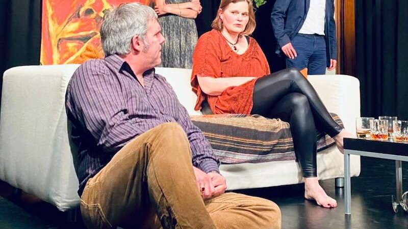 Theaterszene mit vier Personen in einem Wohnzimmer. Eine Person sitzt auf dem Boden, zwei auf dem Sofa, eine steht im Hintergrund. Ein großes Gesichtsgemälde ist sichtbar., © Stadt Sindelfingen