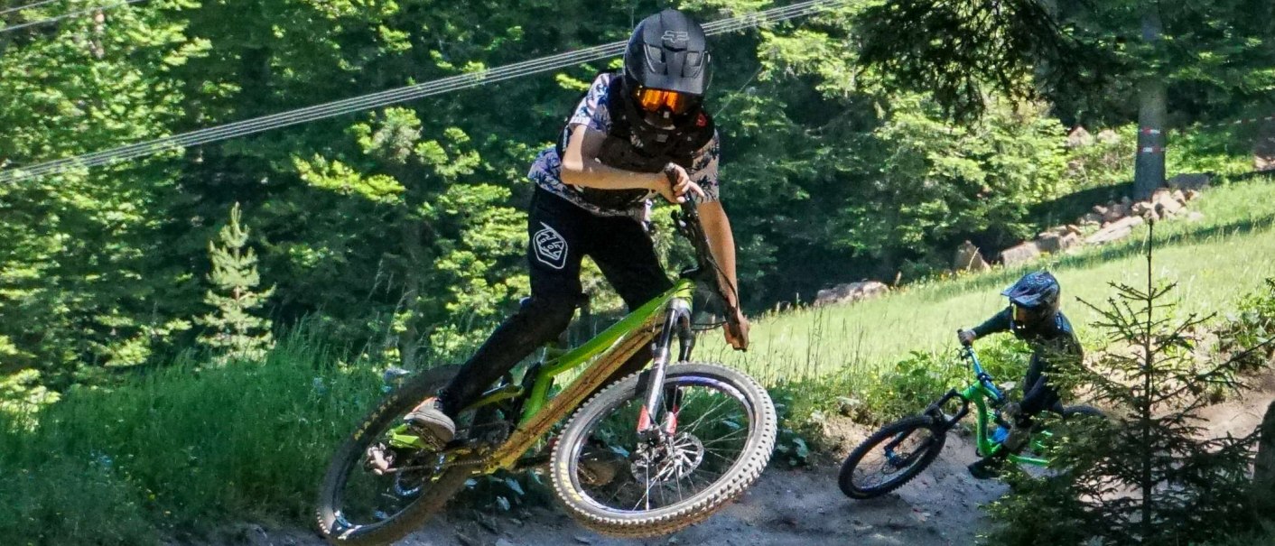 Zwei Mountainbiker mit Helmen fahren auf einem Waldweg im Adventure Bikepark Bad Wildbad. Einer springt, während der andere folgt., © Adventure Sports GmbH