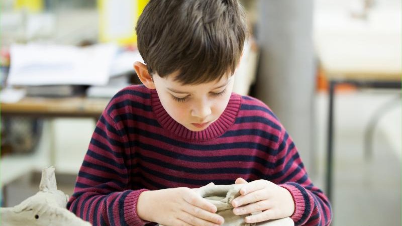 Ein Junge in einem gestreiften Pullover formt konzentriert Ton in einem Workshop. Im Hintergrund sind unscharfe Tische und Materialien zu sehen., © Kunstschule Labyrinth Ein Junge in einem gestreiften Pullover formt konzentriert Ton in einem Workshop. Im Hintergrund sind unscharfe Tische und Materialien zu sehen., © Kunstschule Labyrinth
