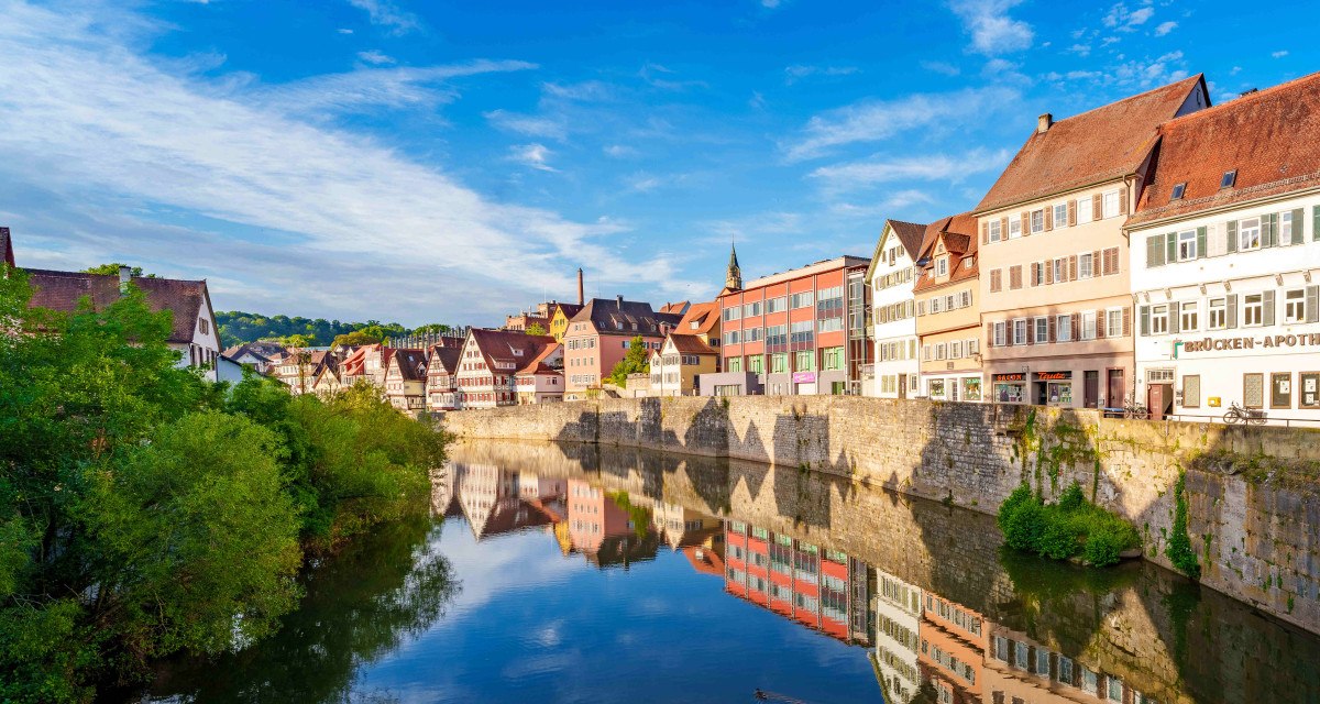 Fachwerkhäuser und moderne Gebäude spiegeln sich im Fluss in Schwäbisch Hall. Klare blaue Himmel und grüne Vegetation rahmen die Szene ein., © Michael Kühneisen