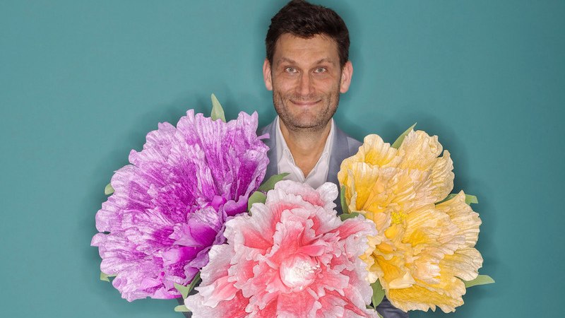 Ein Mann lächelt und hält drei große, bunte Blumen in den Farben Lila, Rosa und Gelb vor einem türkisfarbenen Hintergrund., © Konzertkreis Triangel Compagnia Compostelli e.V. Ein Mann lächelt und hält drei große, bunte Blumen in den Farben Lila, Rosa und Gelb vor einem türkisfarbenen Hintergrund., © Konzertkreis Triangel Compagnia Compostelli e.V.