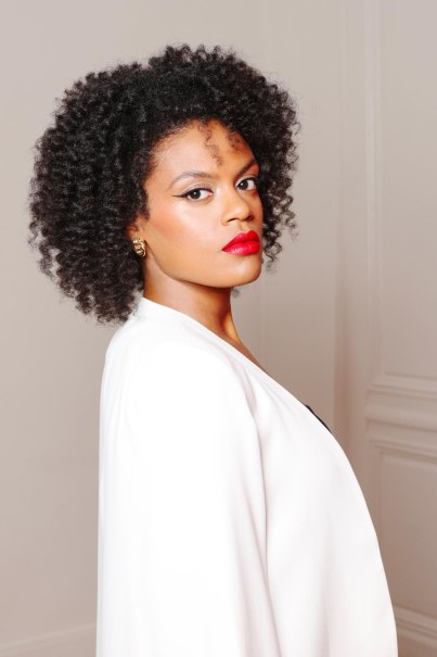 Frau mit lockigem Haar und rotem Lippenstift in weißem Mantel, seitlich blickend., © Ludovic Baron Frau mit lockigem Haar und rotem Lippenstift in weißem Mantel, seitlich blickend., © Ludovic Baron