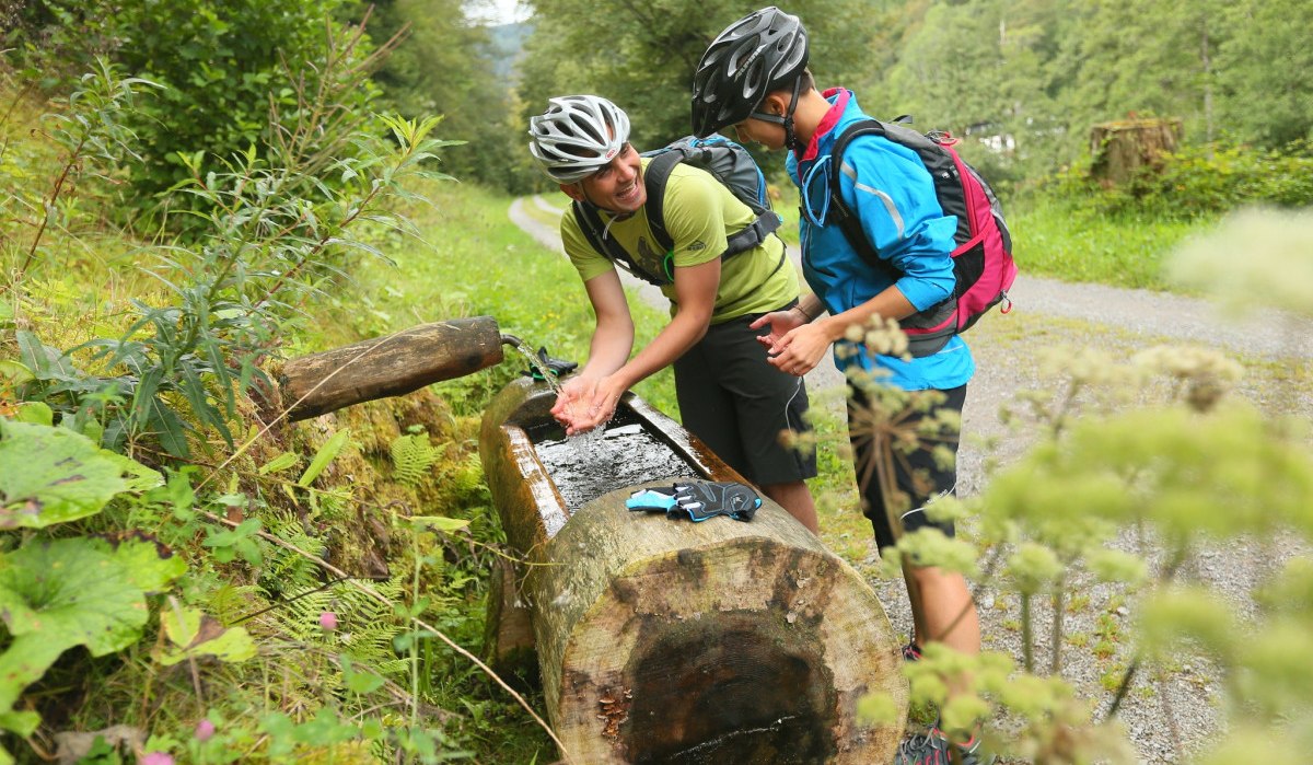Zwei Radfahrer mit Helmen erfrischen sich an einem Holzbrunnen im Wald. Sie lachen und genießen die Pause auf einem Waldweg., © Nördlicher Schwarzwald Zwei Radfahrer mit Helmen erfrischen sich an einem Holzbrunnen im Wald. Sie lachen und genießen die Pause auf einem Waldweg., © Nördlicher Schwarzwald