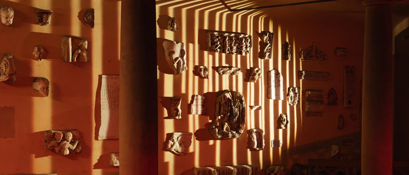 Antike Reliefs und Skulpturen an einer orangefarbenen Wand, beleuchtet von Streifenlicht, das durch eine Säule fällt. Die Szene wirkt warm und atmosphärisch., © Lichtgestalten Antike Reliefs und Skulpturen an einer orangefarbenen Wand, beleuchtet von Streifenlicht, das durch eine Säule fällt. Die Szene wirkt warm und atmosphärisch., © Lichtgestalten