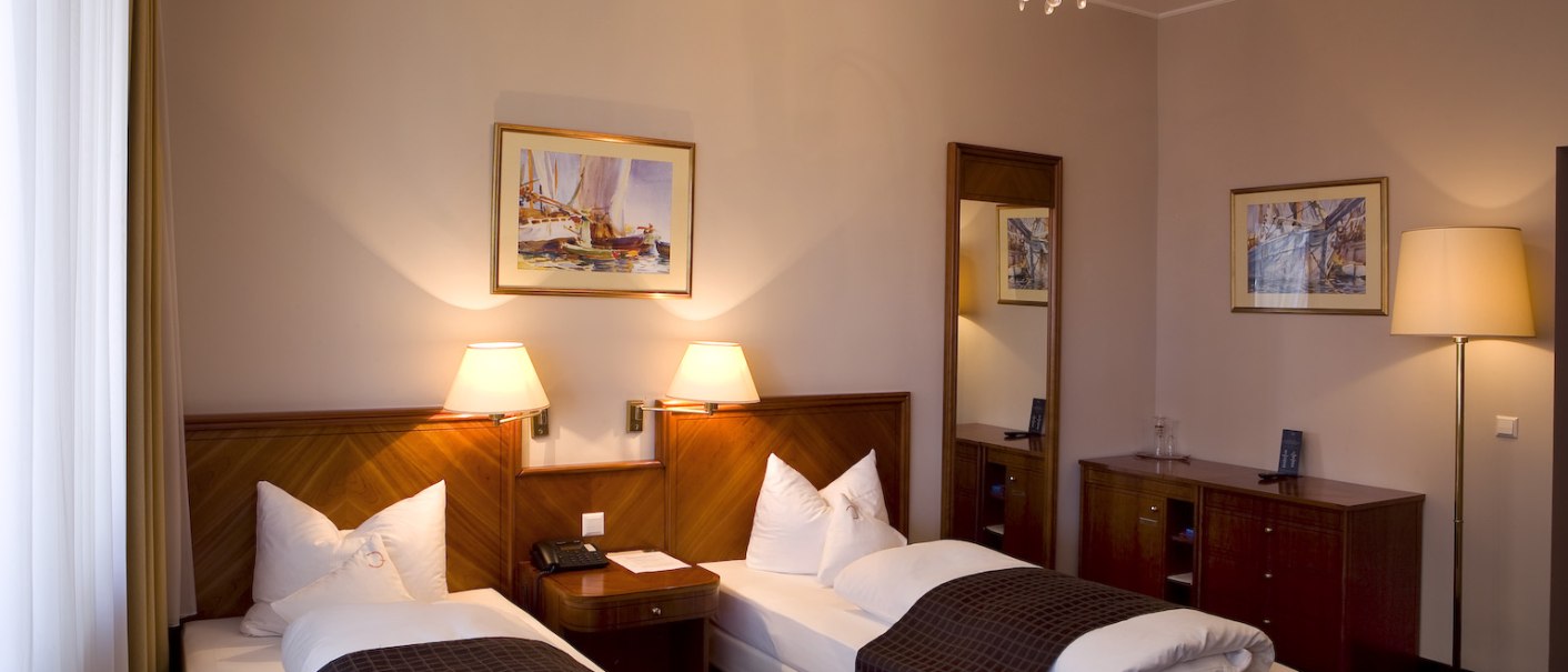 Elegantes Twin-Zimmer mit zwei Einzelbetten, Nachttischlampen, Spiegel, Schrank und Wandbildern. Ein Kronleuchter sorgt für stilvolle Beleuchtung., © Ross Messehotel Elegantes Twin-Zimmer mit zwei Einzelbetten, Nachttischlampen, Spiegel, Schrank und Wandbildern. Ein Kronleuchter sorgt für stilvolle Beleuchtung., © Ross Messehotel