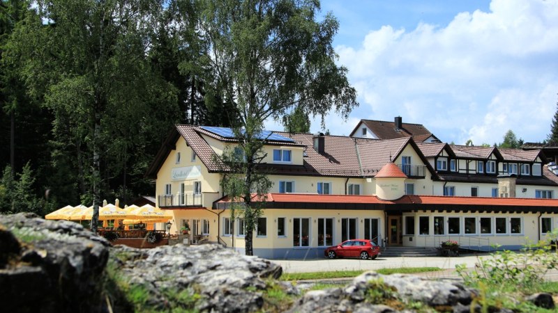 Das Landhotel Wental in Bartholomä, umgeben von Bäumen. Ein rotes Auto steht davor, Sonnenschirme sind aufgestellt. Der Himmel ist blau mit wenigen Wolken., © Landhotel Wental