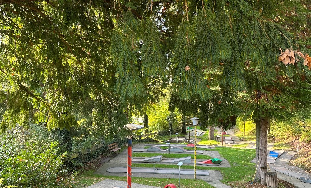 Ein Minigolfplatz im Wald mit mehreren Bahnen und bunten Hindernissen. Die Umgebung ist grün und von Bäumen umgeben., © Ulrike Pfizenmaier Ein Minigolfplatz im Wald mit mehreren Bahnen und bunten Hindernissen. Die Umgebung ist grün und von Bäumen umgeben., © Ulrike Pfizenmaier
