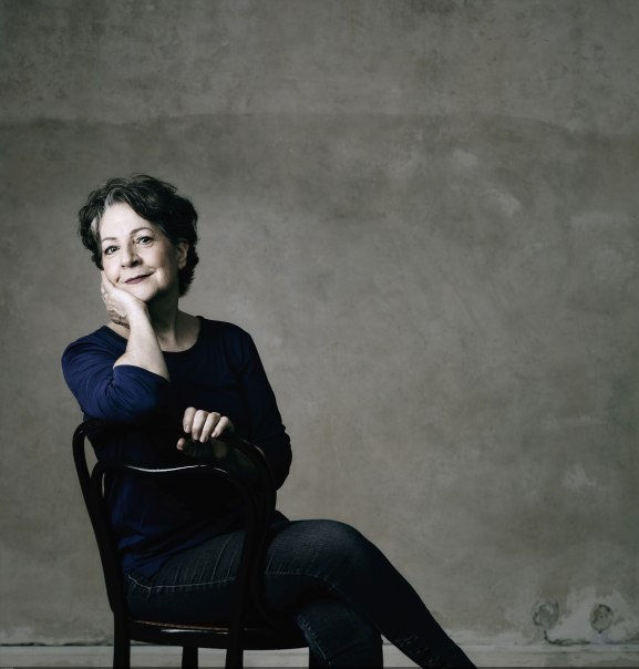 Frau in dunkler Kleidung sitzt lächelnd auf einem Stuhl vor grauer Wand. Frau in dunkler Kleidung sitzt lächelnd auf einem Stuhl vor grauer Wand.