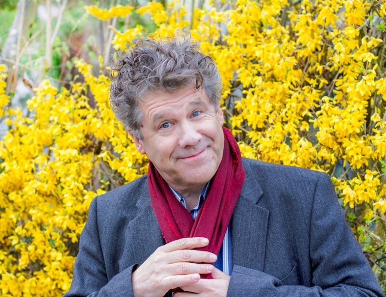 Ein Mann mit lockigem Haar und rotem Schal lächelt vor einem Hintergrund aus gelben Blumen., © Stadt Leinfelden-Echterdingen, Kulturamt