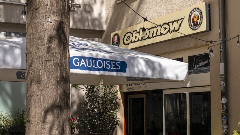 Eingang eines Cafés namens Oblamow mit Gauloises-Sonnenschirm und Pflanzen. Außenbeleuchtung und Schilder von Franziskaner und San Miguel sichtbar., © SMG, Sarah Schmid Eingang eines Cafés namens Oblamow mit Gauloises-Sonnenschirm und Pflanzen. Außenbeleuchtung und Schilder von Franziskaner und San Miguel sichtbar., © SMG, Sarah Schmid