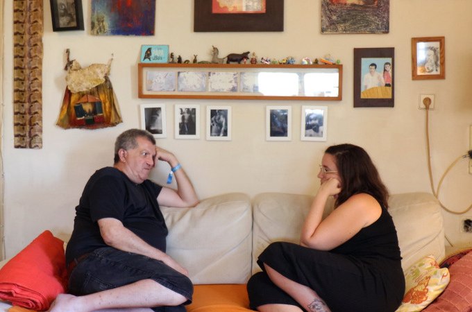 Zwei Personen sitzen auf einem Sofa in einem Wohnzimmer voller Bilder und unterhalten sich entspannt., © Kunsthalle Göppingen Zwei Personen sitzen auf einem Sofa in einem Wohnzimmer voller Bilder und unterhalten sich entspannt., © Kunsthalle Göppingen