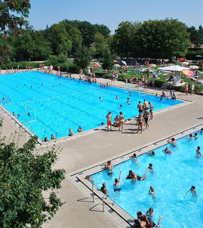 Zwei Schwimmbecken im Waldfreibad Holzgerlingen, umgeben von Bäumen und Liegewiesen. Viele Menschen schwimmen und entspannen sich in der Sonne., © Stadt Holzgerlingen