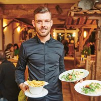 Ein Kellner im Restaurant Himmelreich Schorndorf serviert zwei Teller mit Pasta und eine Schüssel Pommes. Im Hintergrund sitzen Gäste an Holztischen., © Restaurant Himmelreich Schorndorf