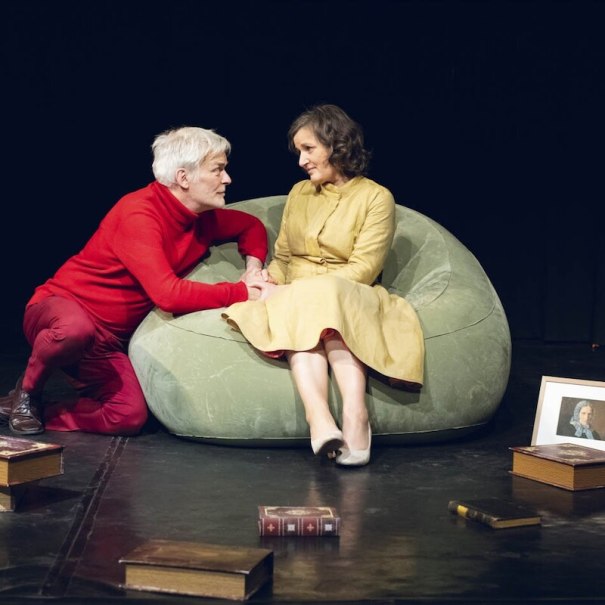 Zwei Personen auf einer Bühne, eine in Rot, die andere in Gelb, sitzen auf einem grünen Sitzsack. Um sie herum liegen Bücher und ein gerahmtes Bild., © Mauerwerk GmbH