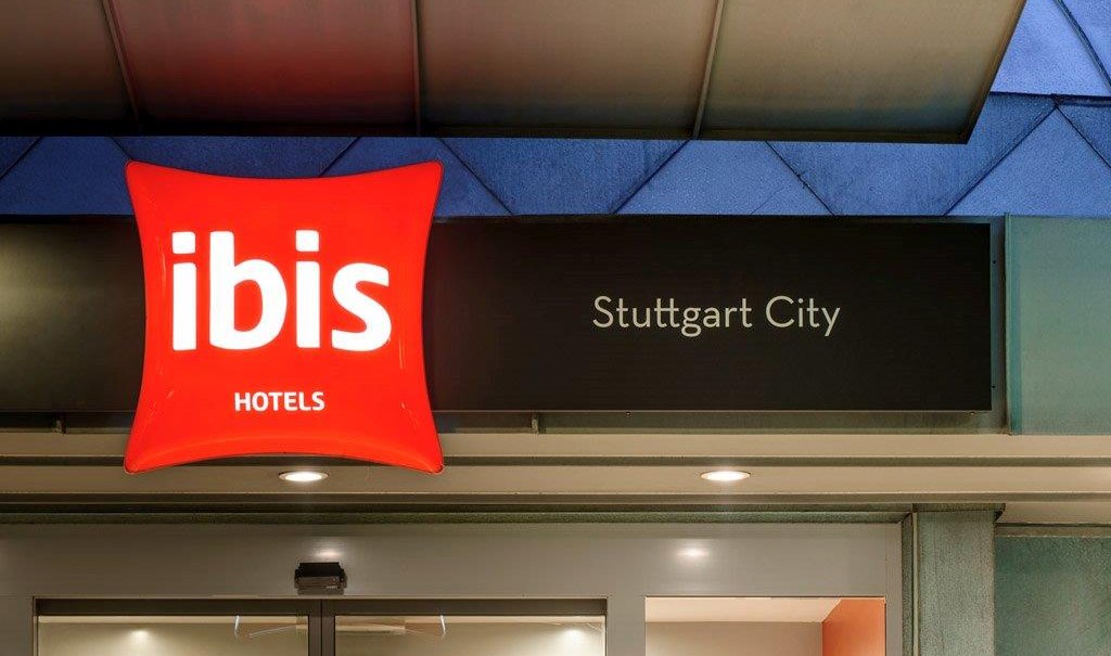 Eingang des Ibis Hotels in Stuttgart City mit leuchtendem roten Logo und schwarzem Schild., © TOMAS