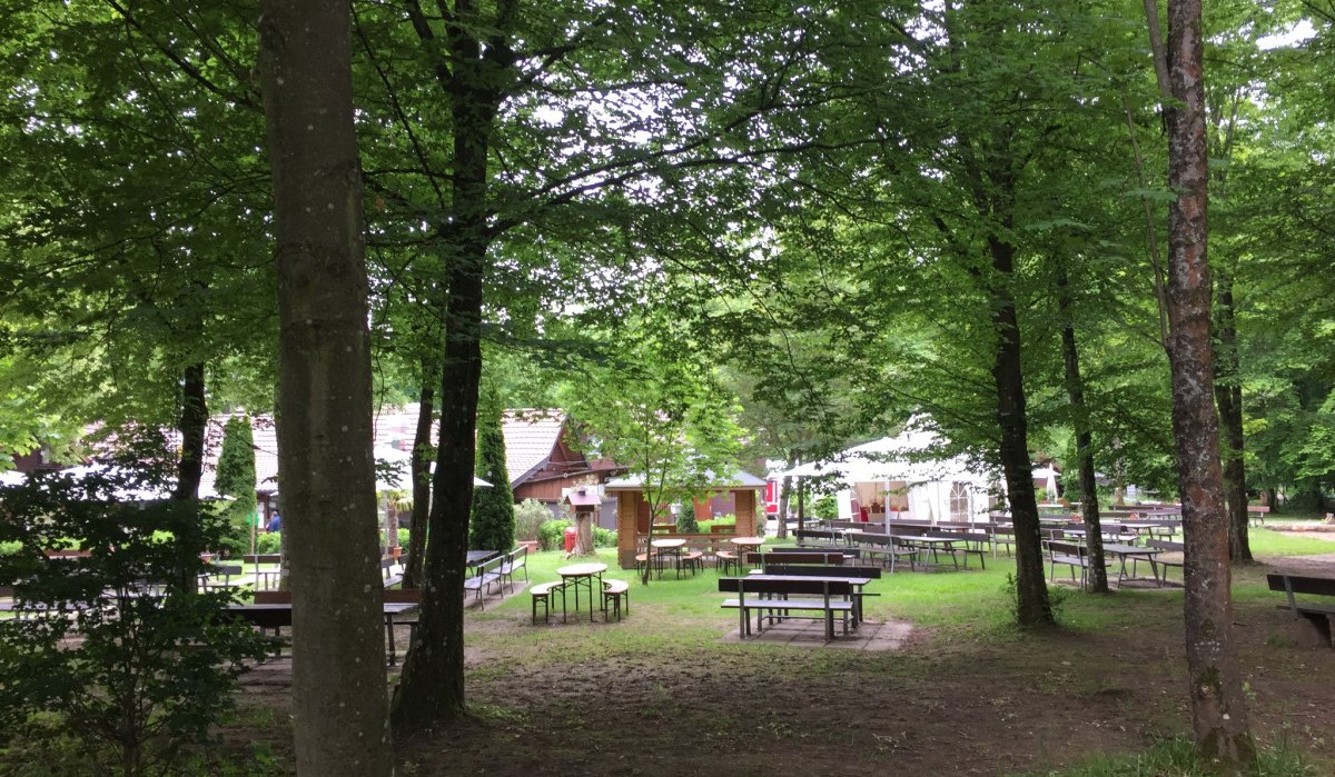 Ein idyllischer Biergarten im Wald mit leeren Holztischen und B&auml;nken, umgeben von gr&uuml;nen B&auml;umen und einem kleinen Geb&auml;ude im Hintergrund., &copy; www.pro-cycl.de