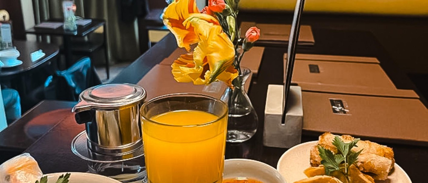 Asiatische Gerichte auf einem Tisch: Frühlingsrollen, Dumplings, Reis, Gemüse und ein Glas Orangensaft. Im Hintergrund ein gemütliches Restaurantambiente., © by Canto