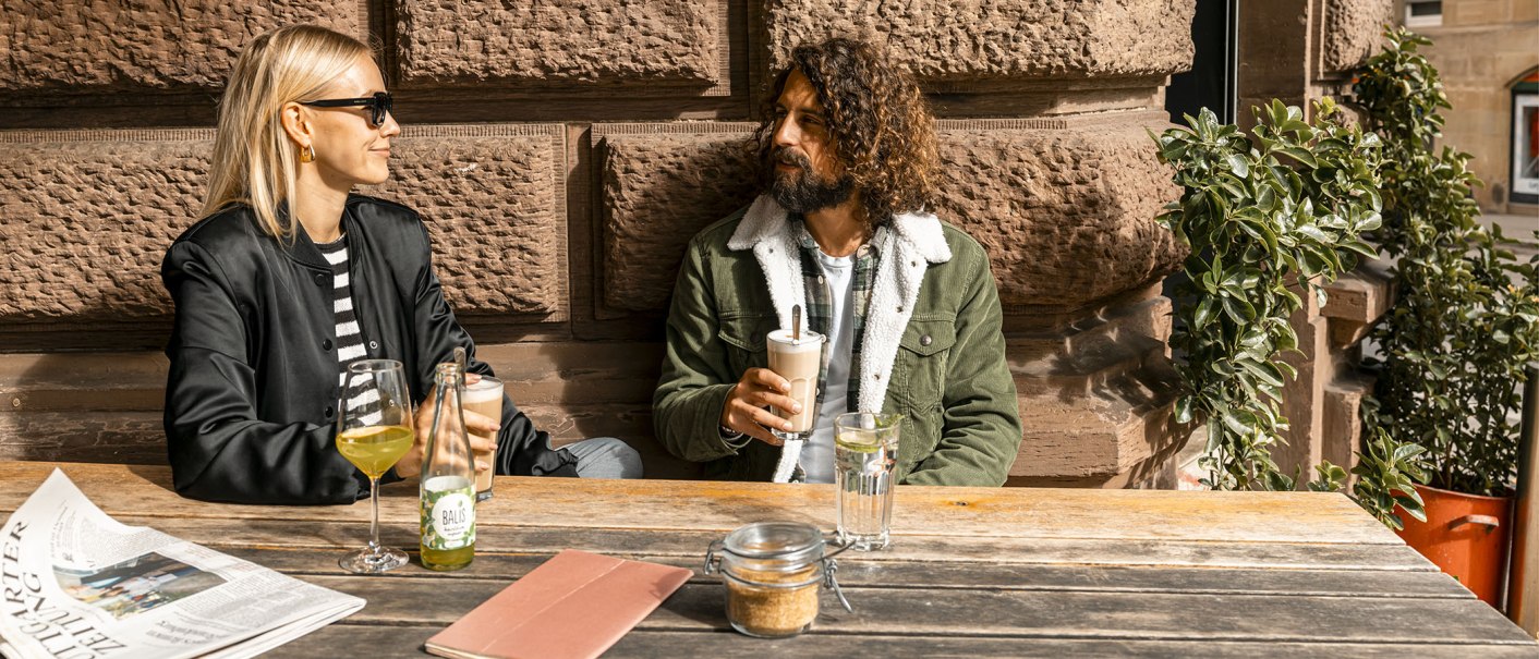 Zwei Personen sitzen an einem Holztisch vor einer Steinmauer, trinken Getränke und unterhalten sich. Eine Zeitung liegt auf dem Tisch., © SMG, Sarah Schmid