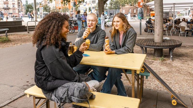 Drei Personen sitzen an einem gelben Tisch im Freien und essen Eis. Im Hintergrund sind B&auml;ume, Fahrr&auml;der und ein Caf&eacute; zu sehen., &copy; Stuttgart Marketing GmbH, Sarah Schmid
