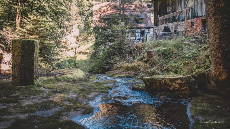 Ein Bach fließt durch einen Wald, vorbei an einem alten Fachwerkhaus. Moosbewachsene Steine und Bäume umgeben die Szene., © Stadt Welzheim Ein Bach fließt durch einen Wald, vorbei an einem alten Fachwerkhaus. Moosbewachsene Steine und Bäume umgeben die Szene., © Stadt Welzheim