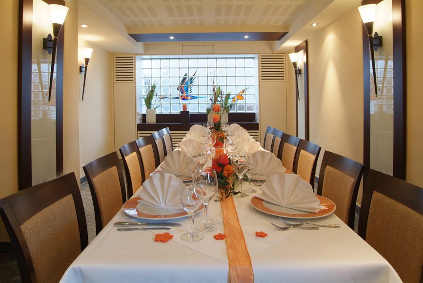Eleganter Raum mit langem Tisch, gedeckt mit weißen Servietten, Gläsern und orangefarbener Dekoration. Wandlampen und Glasfenster im Hintergrund., © Römerhof Hotelbetriebs GmbH