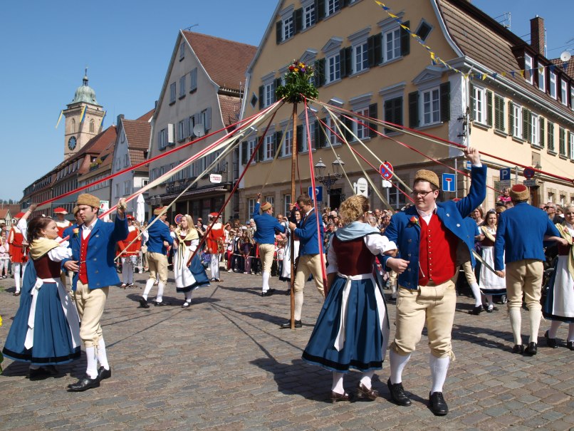 Menschen in traditioneller Kleidung tanzen um einen Maibaum auf einem gepflasterten Platz in Nürtingen. Im Hintergrund sind historische Gebäude zu sehen., © Stadt Nürtingen