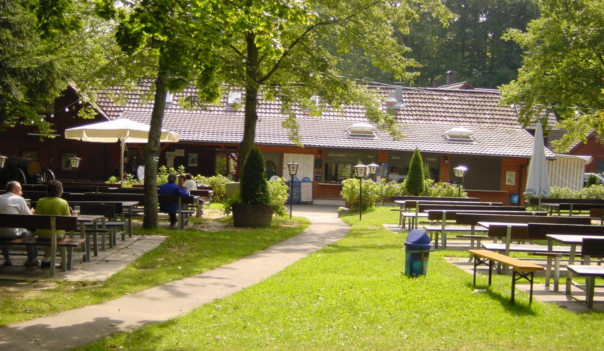 Ein Biergarten mit Holzbänken und Tischen im Freien, umgeben von Bäumen. Im Hintergrund ein Gebäude mit Ziegeldach., © Naturpark Schönbuch