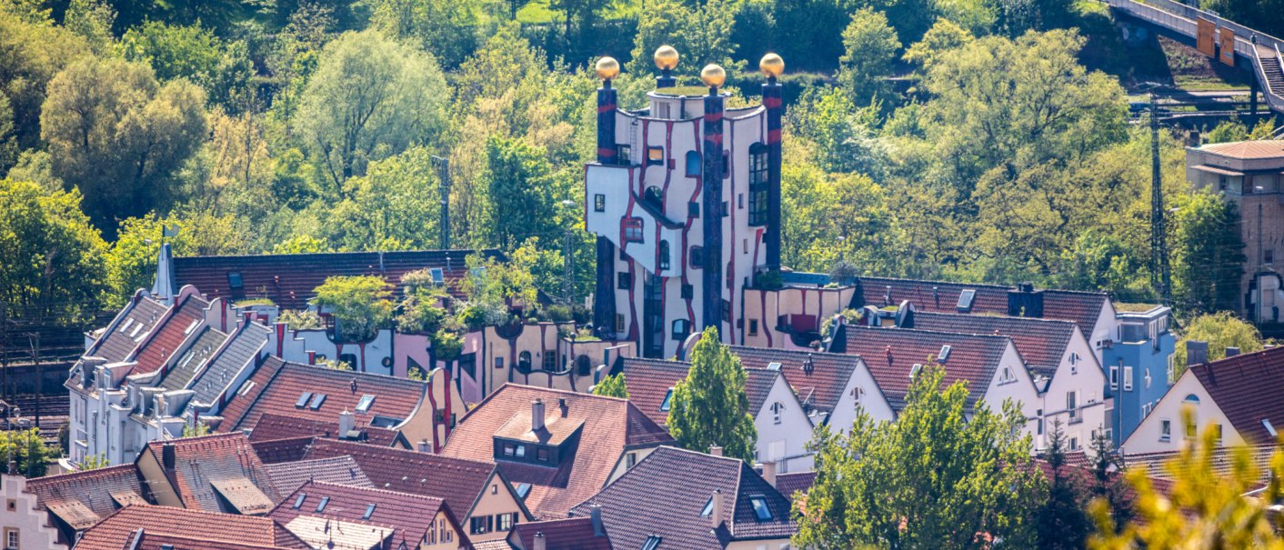Das Hundertwasser-Wohnhaus in Plochingen zeigt bunte Fassaden und goldene Kuppeln, umgeben von Bäumen und traditionellen Häusern., © SMG Achim Mende Das Hundertwasser-Wohnhaus in Plochingen zeigt bunte Fassaden und goldene Kuppeln, umgeben von Bäumen und traditionellen Häusern., © SMG Achim Mende
