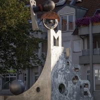 Der Katz- und Mausbrunnen in Bottwartal zeigt eine moderne Skulptur mit Buchstaben und geometrischen Formen vor einem Gebäude mit roten Dachziegeln., © Stuttgart Marketing