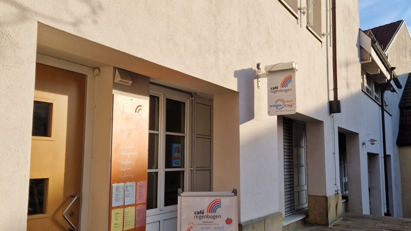 Das Bild zeigt die Außenansicht des Café Regenbogen mit einem Schild und einem Aufsteller vor der Eingangstür. Die Fassade ist hell und die Straße gepflastert., © StadtNuertingen Das Bild zeigt die Außenansicht des Café Regenbogen mit einem Schild und einem Aufsteller vor der Eingangstür. Die Fassade ist hell und die Straße gepflastert., © StadtNuertingen