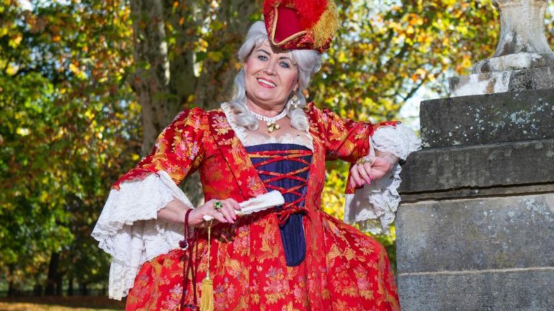 Eine Person in einem aufwendigen, roten historischen Kostüm posiert lächelnd neben einer Steinsäule im Freien. Herbstliche Bäume im Hintergrund., © © T&E, Reiner Pfisterer Eine Person in einem aufwendigen, roten historischen Kostüm posiert lächelnd neben einer Steinsäule im Freien. Herbstliche Bäume im Hintergrund., © © T&E, Reiner Pfisterer