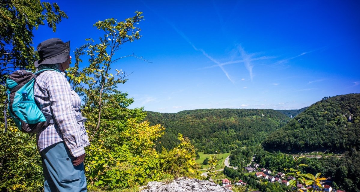 Eine Person mit Rucksack steht auf einem Hügel und blickt auf ein bewaldetes Tal mit einem Dorf. Der Himmel ist klar und blau., © Stadt Geislingen an der Steige Eine Person mit Rucksack steht auf einem Hügel und blickt auf ein bewaldetes Tal mit einem Dorf. Der Himmel ist klar und blau., © Stadt Geislingen an der Steige