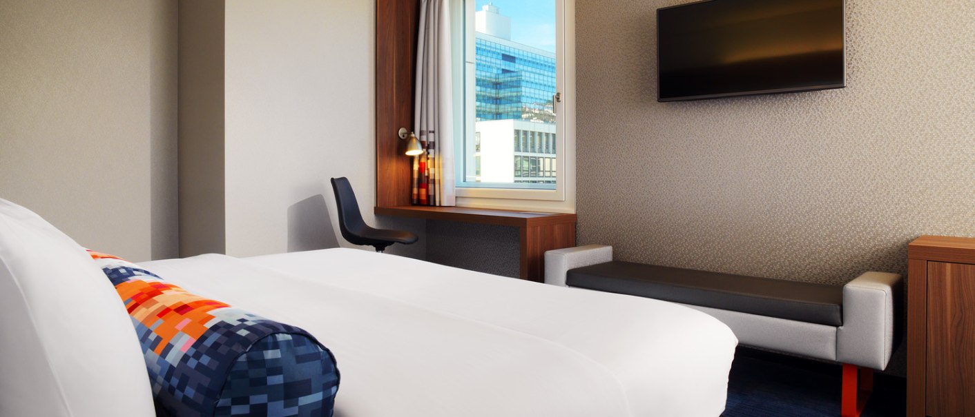 Ein modernes Hotelzimmer mit einem gro&szlig;en Bett, einem Schreibtisch am Fenster und einem Flachbildfernseher an der Wand., &copy; Aloft Stuttgart